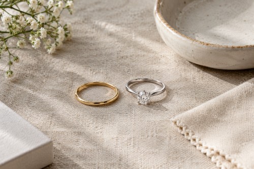 Slim promise ring next to a diamond solitaire engagement ring on linen, showing the size and design difference